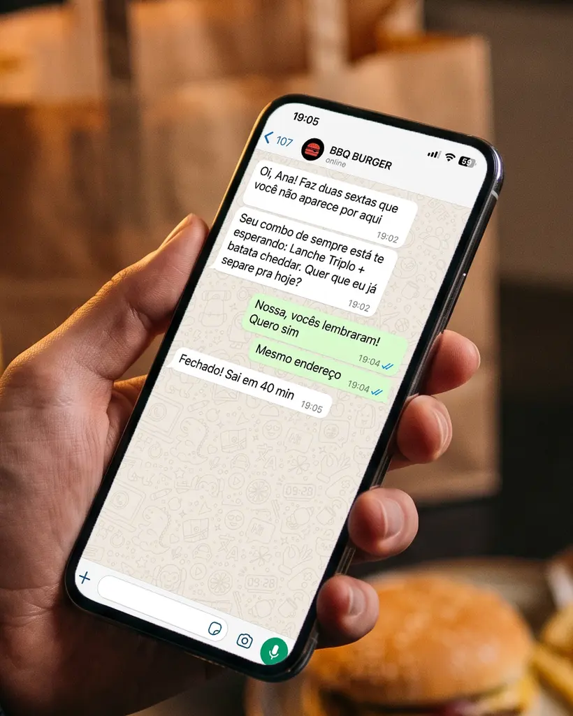 Mão segurando celular com conversa no WhatsApp em que o delivery chama a cliente de volta para o combo de sempre, com hambúrguer e batata desfocados ao fundo